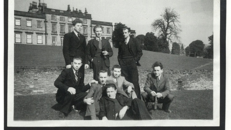 A black and white photo shows a group of eight men dressed in suits, posing outdoors. Five are standing behind three who are seated or kneeling in front. Behind them is a large historic building, likely Cannon Hall, with expansive grounds and a tree.